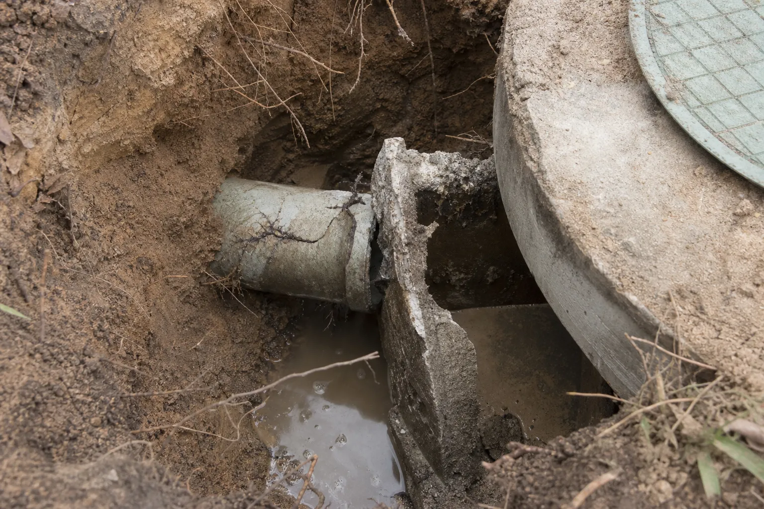 Close-up into a small excavation at a tank inlet: a cracked root-invaded inlet pipe and a broken concrete baffle